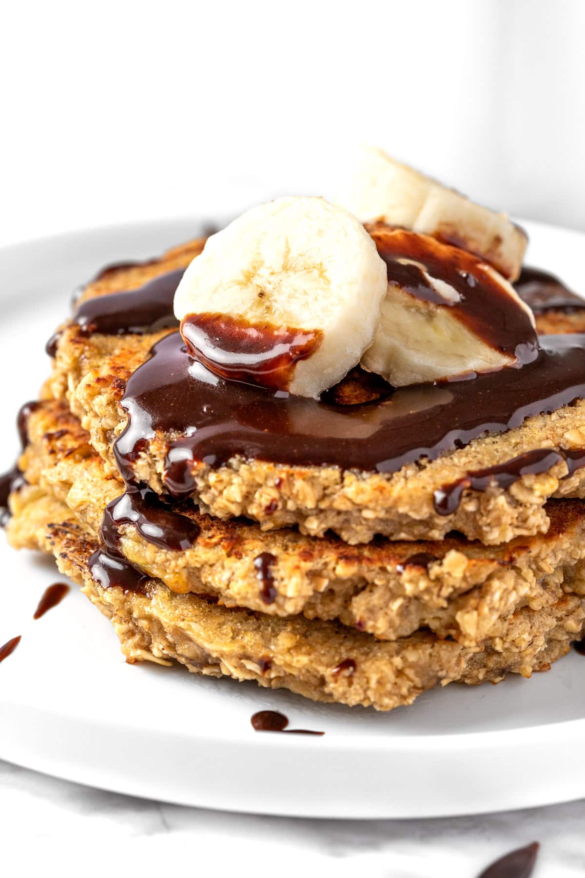 A stack of banana oat pancakes topped with banana slices and chocolate sauce.