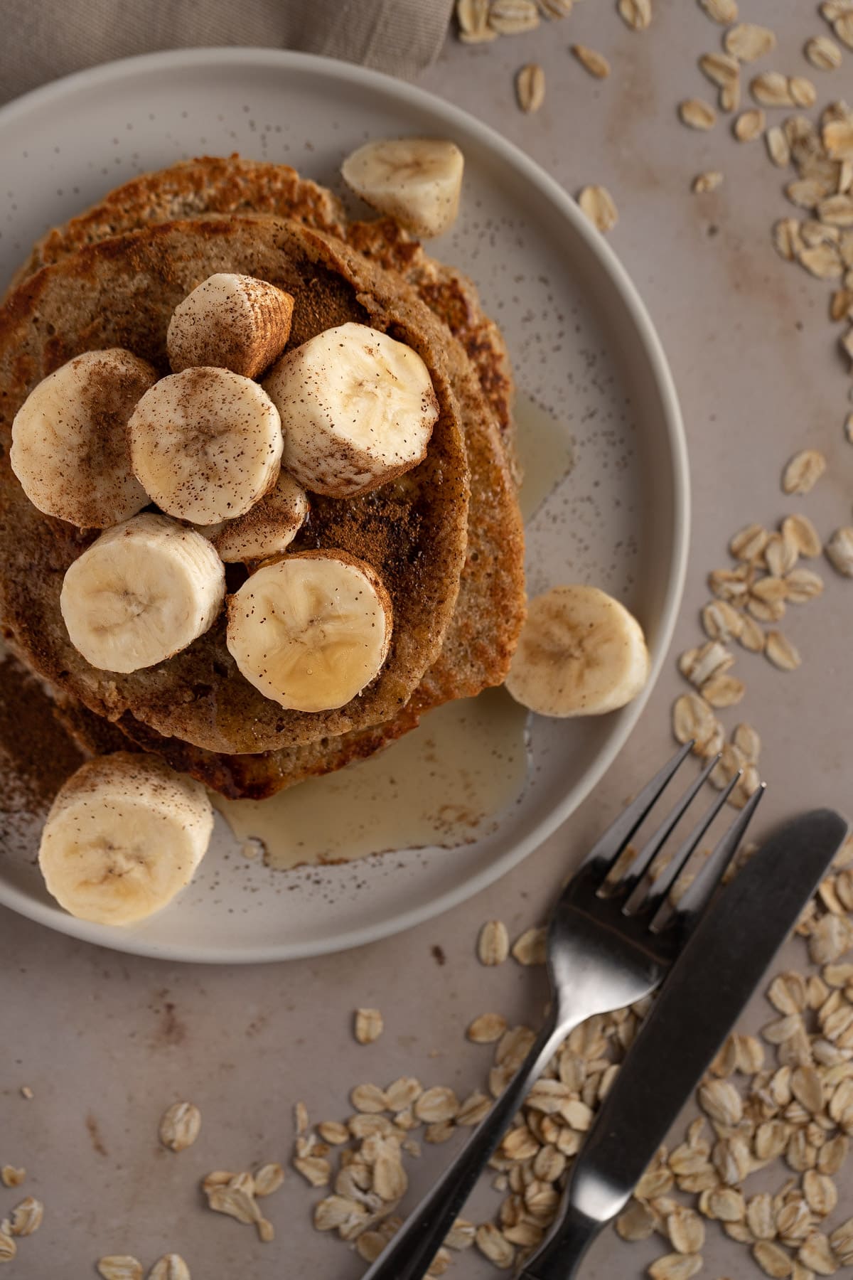 Overhead view of a stack of pancakes topped with banana slices, on a warm marble table covered with scattered oats.