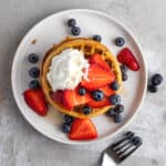 An overhead view of air fryer eggo waffles stacked on a round white plate, topped with strawberries and blueberries.
