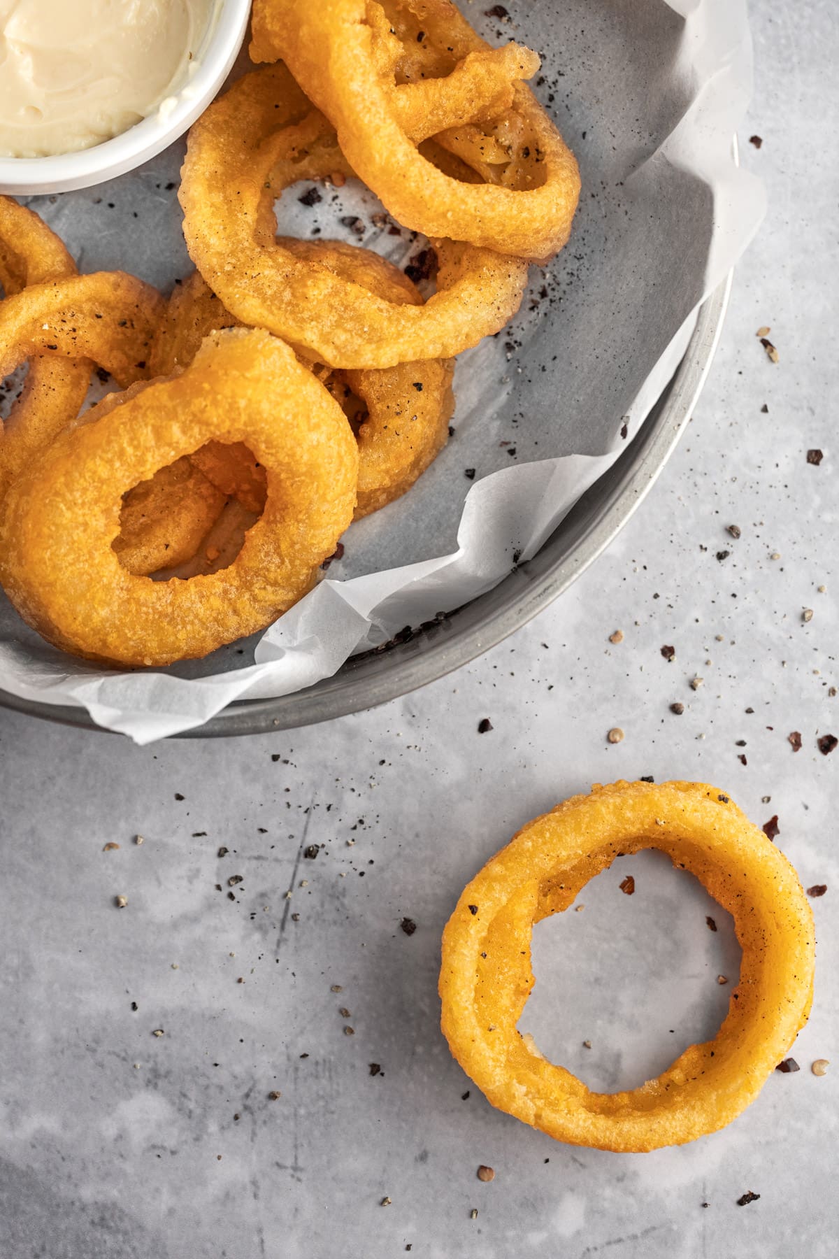 Onion rings in a round metal tray on a grey marble table with one onion ring on the table.