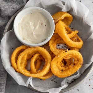 Air fryer frozen onion rings in a round metal tin lined with parchment paper next to a ramekin of mayo dipping sauce.
