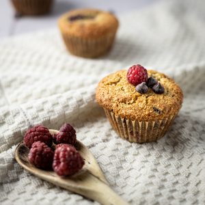 Three almond flour raspberry muffins in a trail, with a wooden spoon full of raspberries in the front