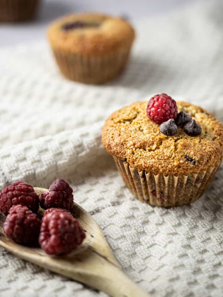 Three almond flour raspberry muffins in a trail, with a wooden spoon full of raspberries in the front