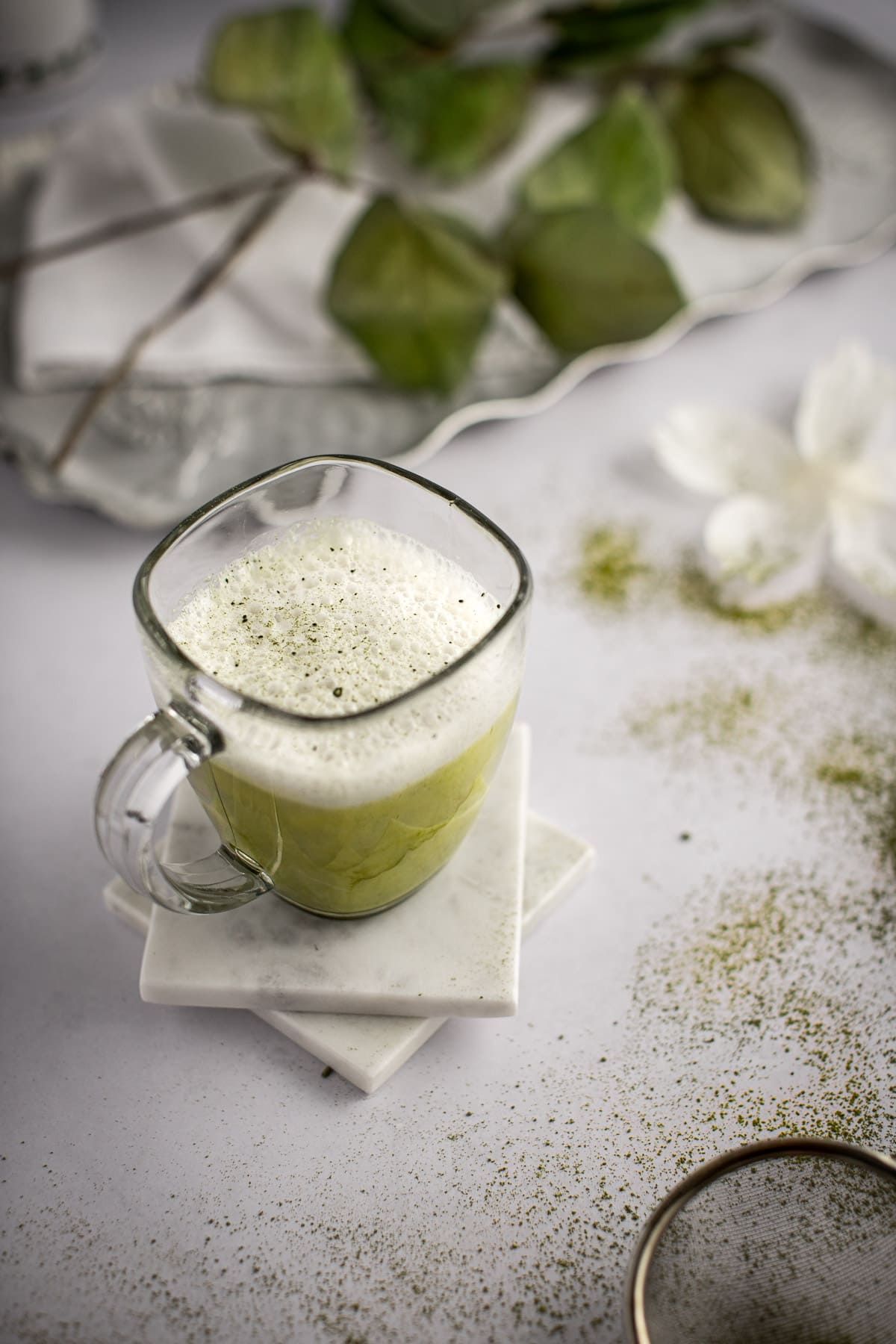 Matcha almond milk latte on two white marble coaster, on a light grey marble table, with white flowers and matcha powder scattered around.