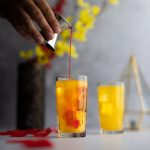 Grenadine being poured into an amaretto sunrise cocktail, with rose petals scattered around and red and yellow flowers in the background.