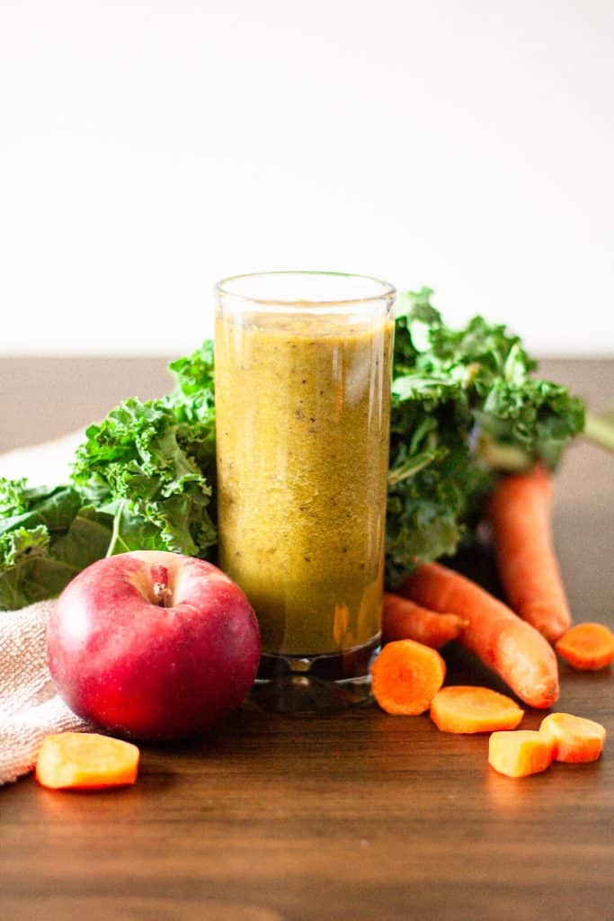 Tall glass of carrot, apple, kale and ginger smoothie with kale leaves, carrots pieces and an apple around the glass.