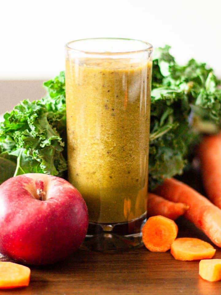 Tall glass of carrot, apple, kale and ginger smoothie with kale leaves, carrots pieces and an apple around the glass.
