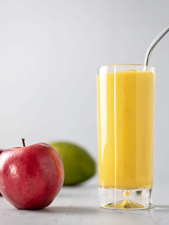 A tall glass of apple mango smoothie with a metal straw sticking out, and a couple apples and mango on the table beside it