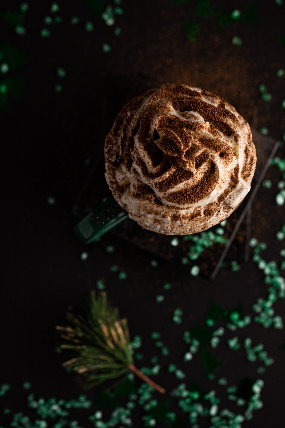 Overhead view of hot chocolate topped with whipped cream and sprinkled cocoa powder, on a black background with green sparkles.