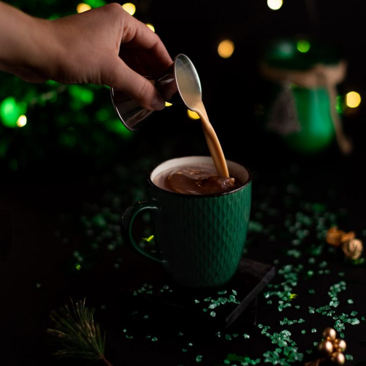 A shot of baileys being poured into the hot chocolate in a green mug with christmas lights in the background.