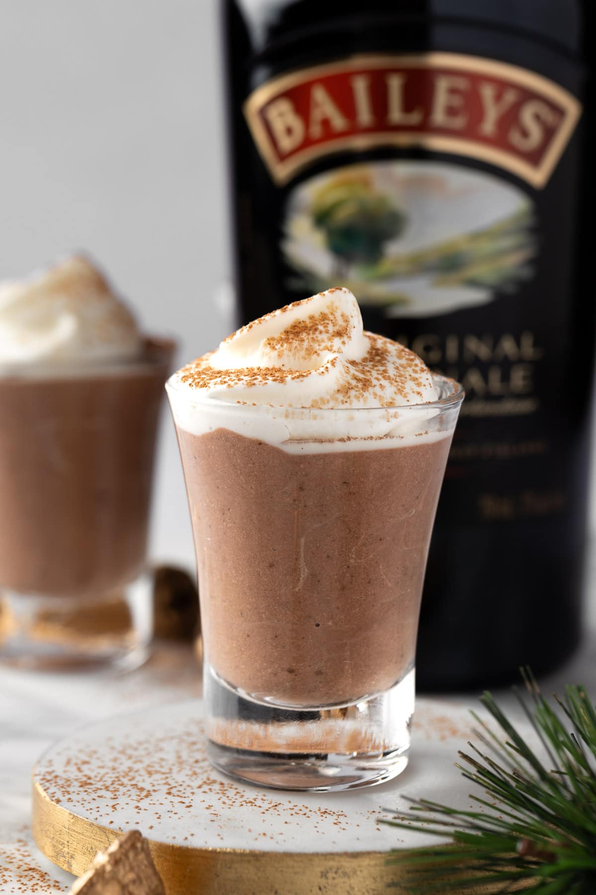 Baileys Pudding Shots topped with whipped cream and a bottle of Baileys in the background.