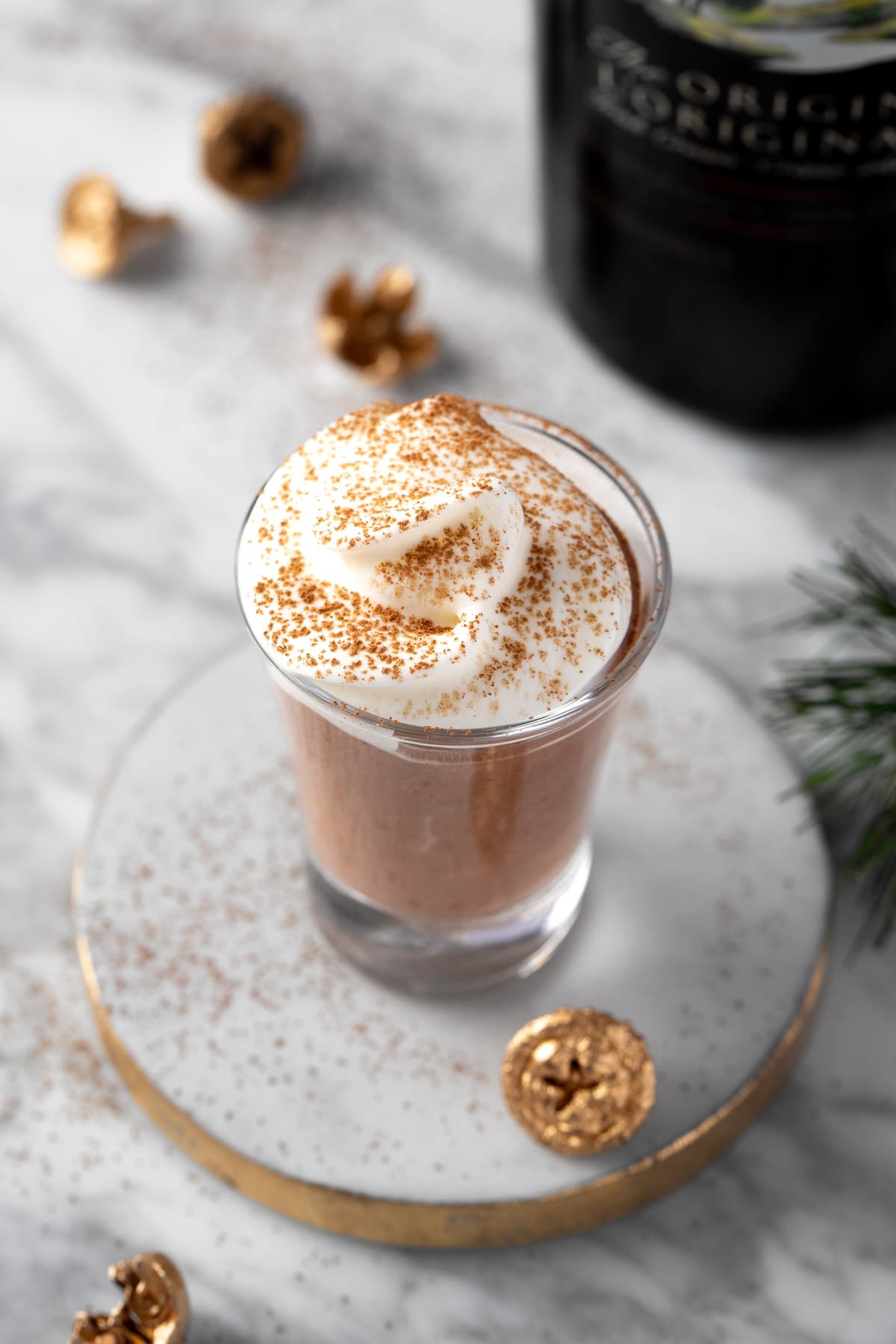 A Baileys Pudding Shot topped with whipped cream, on a white and gold coaster.