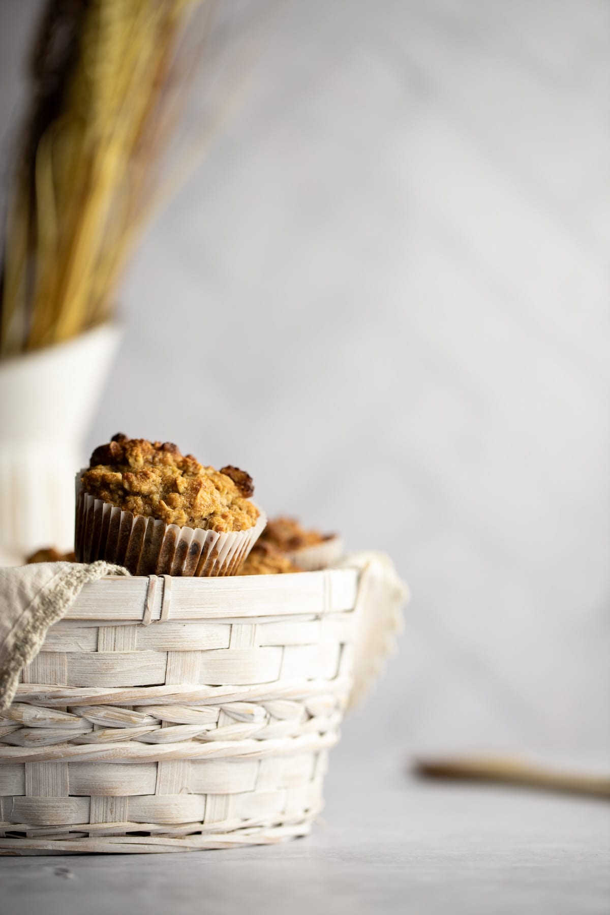 Banana almond flour muffins overflowing from a basket, with a white jug with wheat in it in the background.