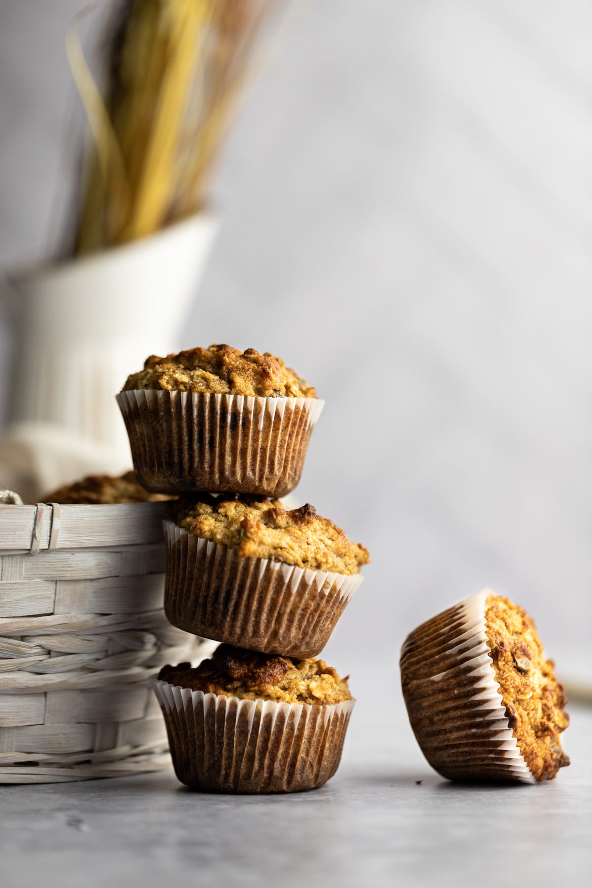 A stack of three almond flour banana muffins leaning against a basket, with one muffin tipped on its side lying beside the stack.