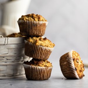A stack of banana almond flour muffins leaning against a white basket, one muffin lying on its side beside the other muffins.