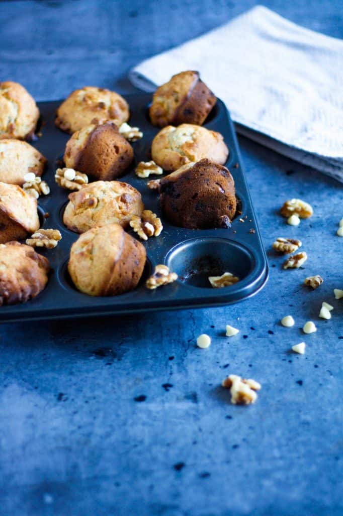 a tin filled with banana white chocolate muffins, scattered with white chocolate chips and walnuts