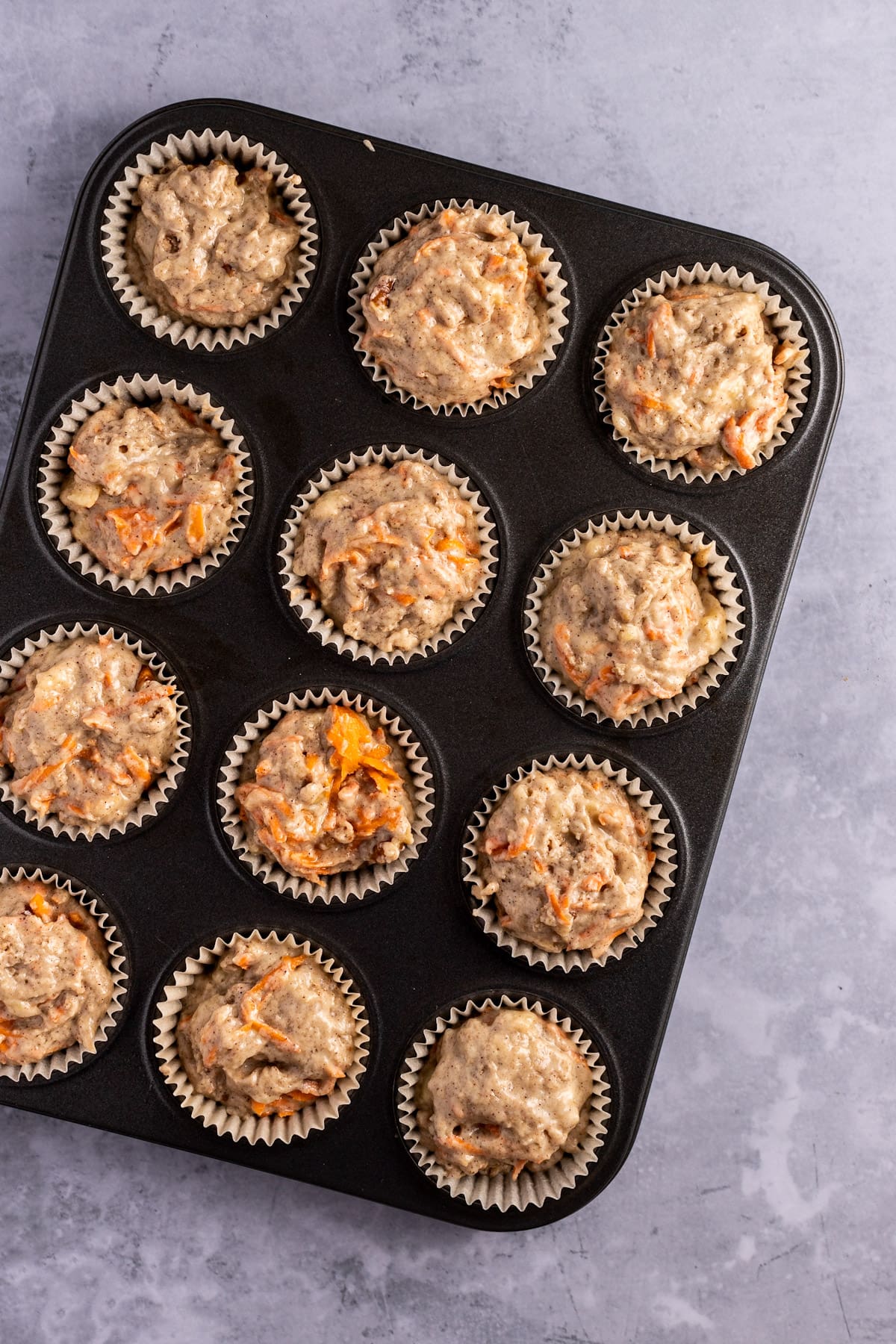 A muffin tray on a grey background, filled with muffin liners that have been filled with muffin batter.