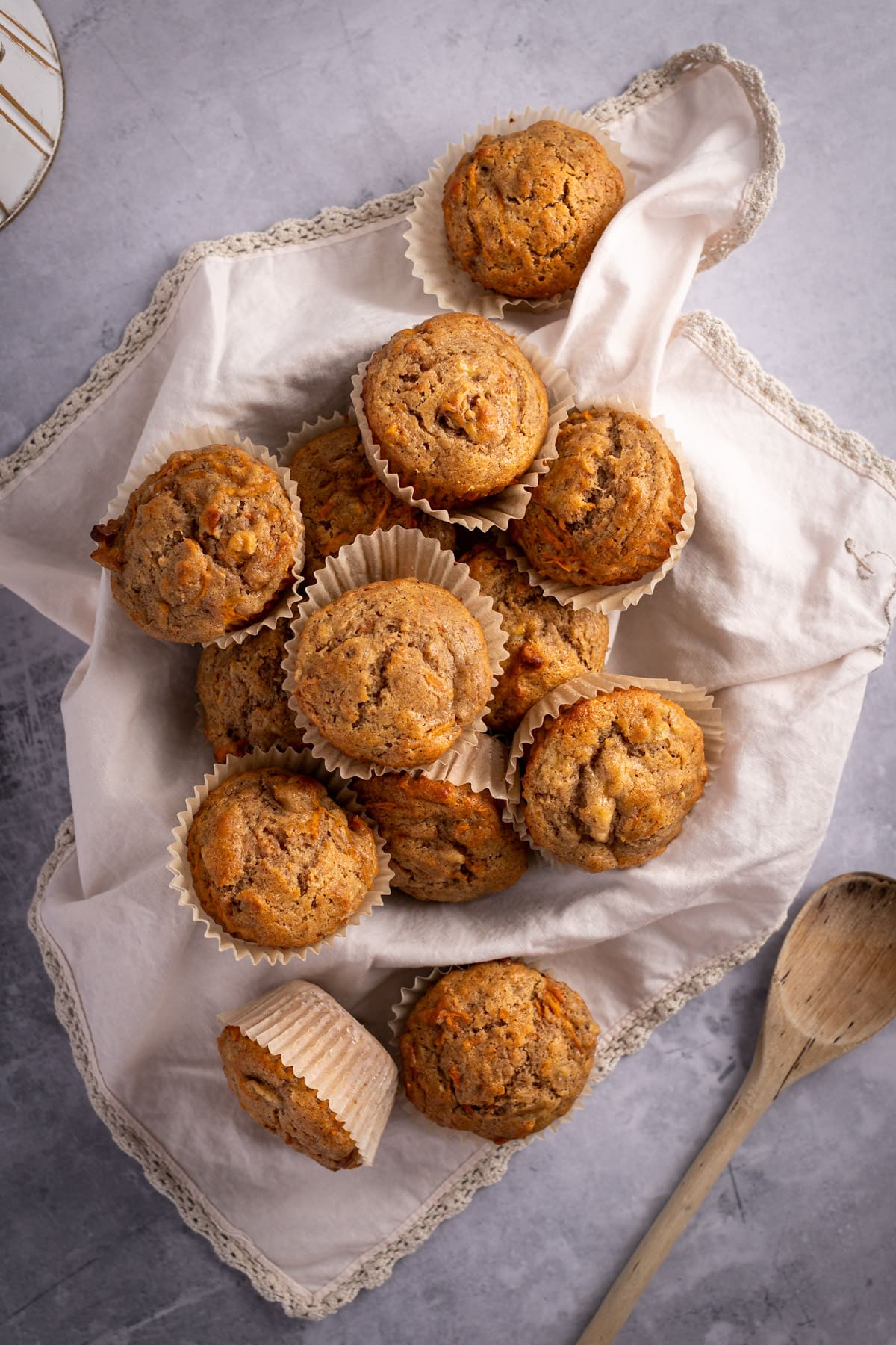 Banana carrot muffins scattered over a light pink cloth, next to a wooden spoon, on a dark grey table.