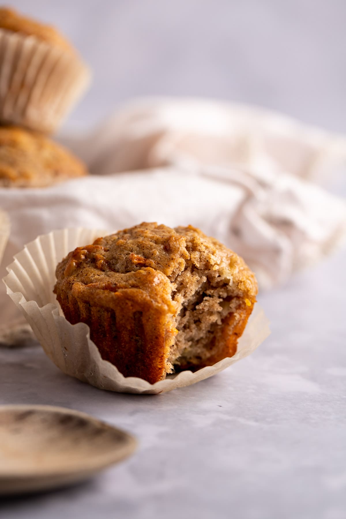 A single banana carrot muffin with a bite missing, with its muffin liner peeled away, sitting on a grey table.