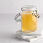 A jar of yellow banana syrup sitting on three white square marble coasters, on a white background.