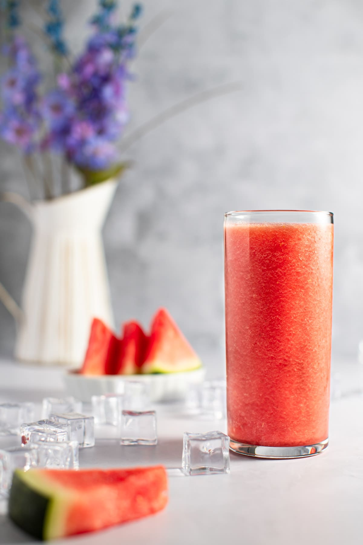 A tall glass of banana watermelon smoothie, next to ice cubes and slices of watermelon, with purple flowers in the background.