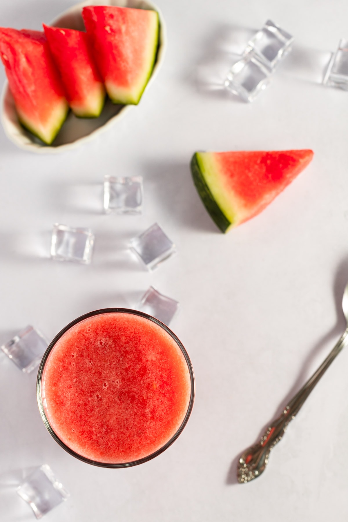 Overhead photo of smoothie, surrounded by a metal spoon and watermelon slices, on a white background.