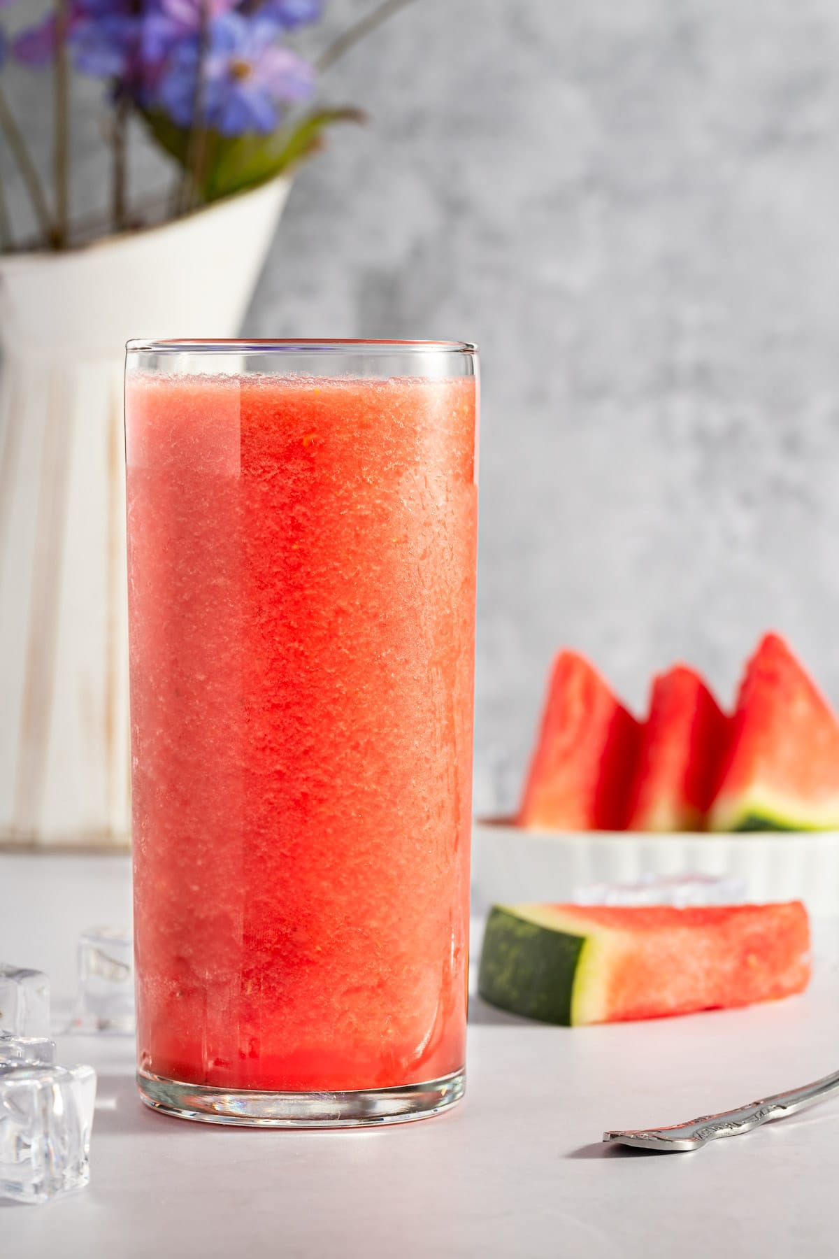 Up close photo of watermelon and banana smoothie, next to slices of watermelon, ice cubes, and a vase of flowers in the background.