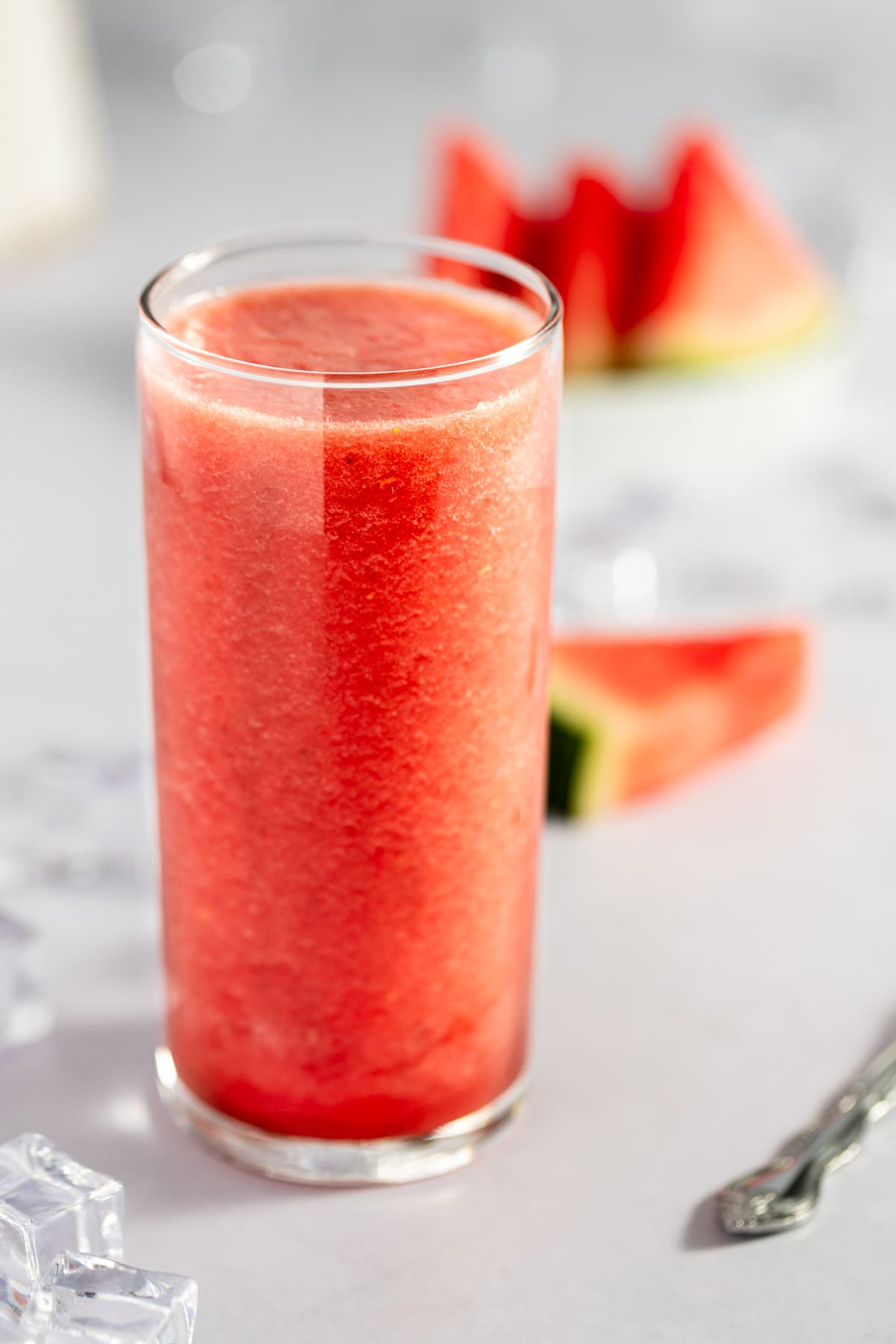 Tall glass of redish-pink watermelon smoothie, with slices of watermelon in the background.