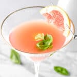 A basil grapefruit martini garnished with a slice of grapefruit, on a white marble table.