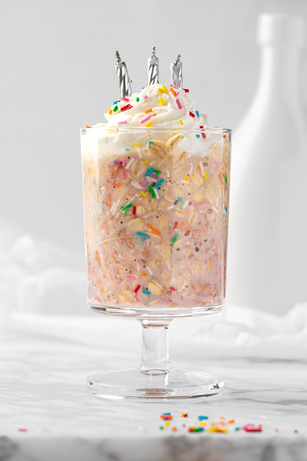 Eye level view of birthday cake overnight oats, topped with whipped cream and sprinkles, on a white background.