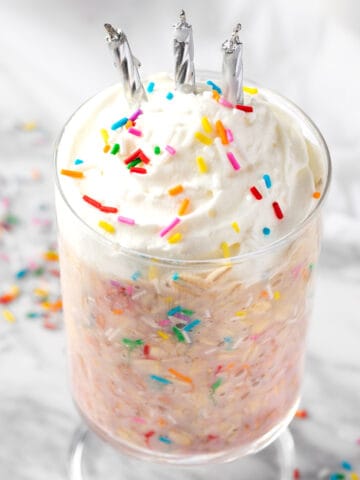 A glass of birthday cake overnight oats topped with whipped cream, sprinkles and candles.