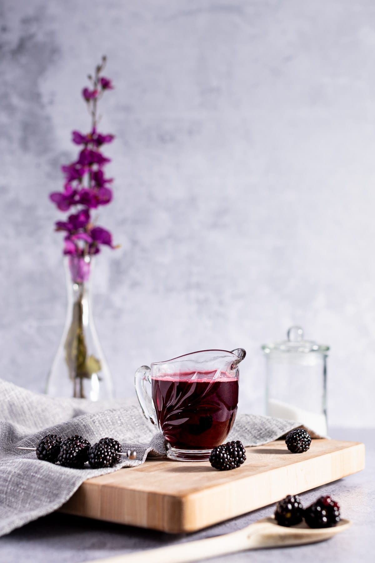 Glass container of simple syrup, sitting on a wooden board, next to fresh blackberries, with a glass vase filled with purple flowers in the background.