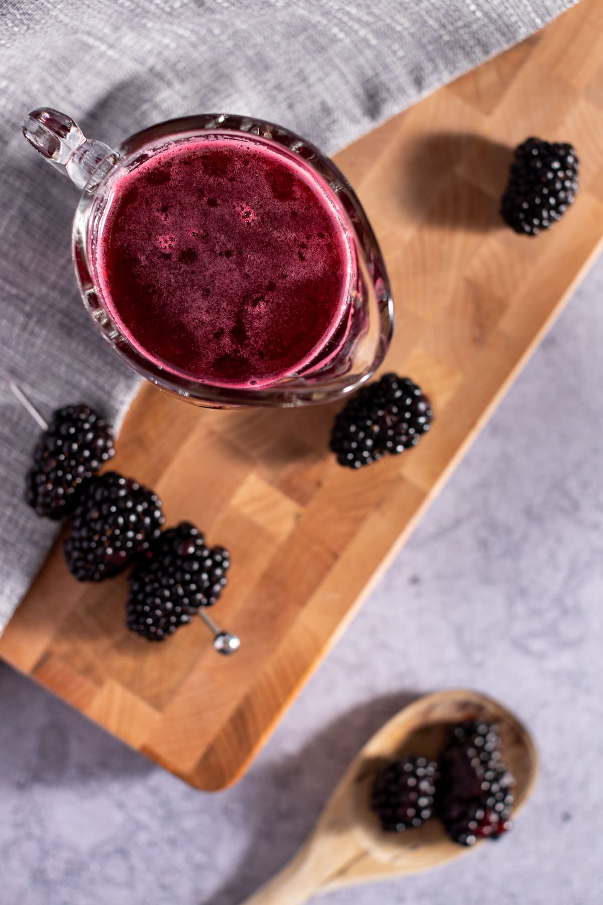 Overhead photo of syrup in a glass container, on a wooden cutting board, next to a wooden spoon holding fresh blackberries.