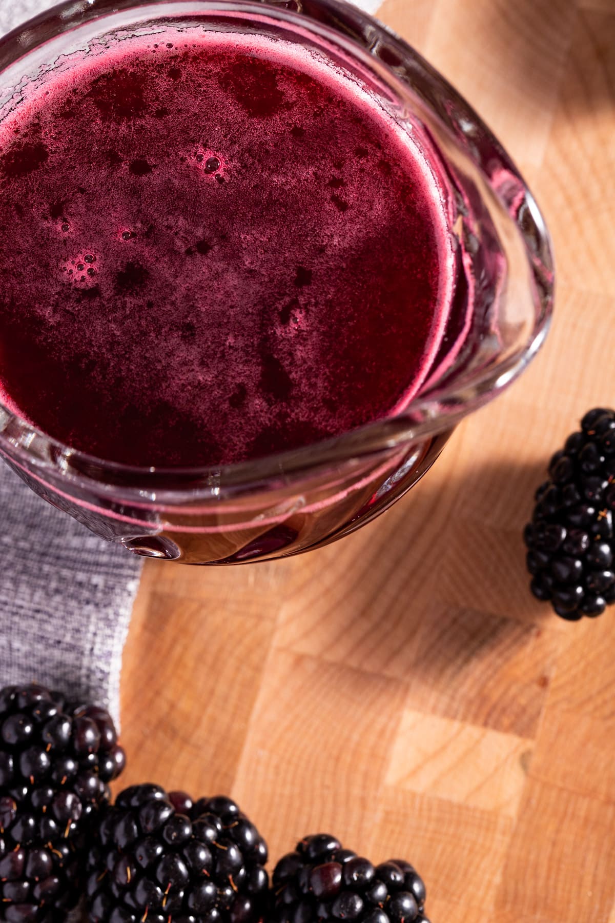 Overhead view of blackberry syrup, next to blackberries, on a wooden cutting board.