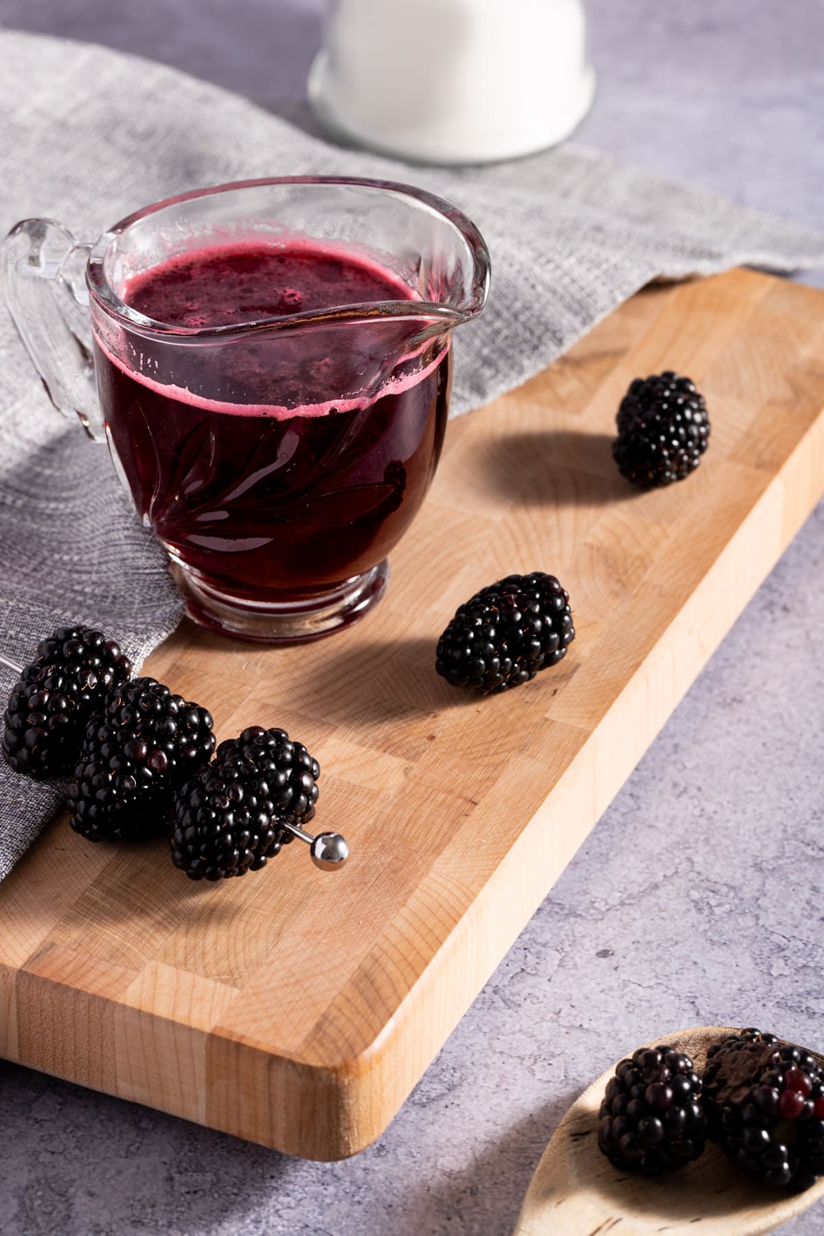 Blackberry simple syrup in a glass container on a wooden cutting board, next to fresh blackberries and a grey linen napkin.