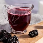 Blackberry simple syrup in a small glass container, next to fresh blackberries, on a wooden board with a grey napkin in the background.