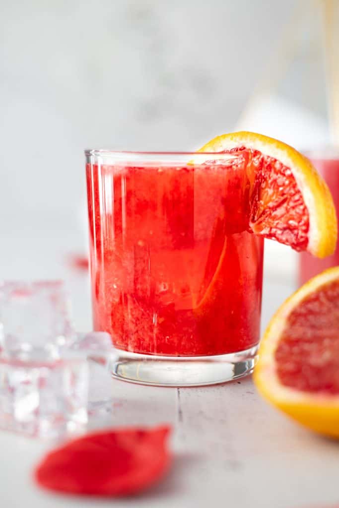 Close up eye level view of a glass of blood orange whiskey sour with ice cubes and rose petals in front