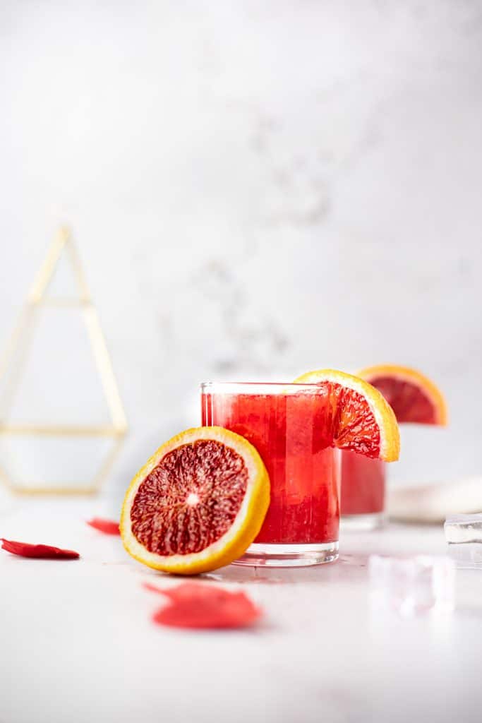 A glass of blood orange whiskey sour with half a blood orange leaning against the glass and rose petals scattered on the table