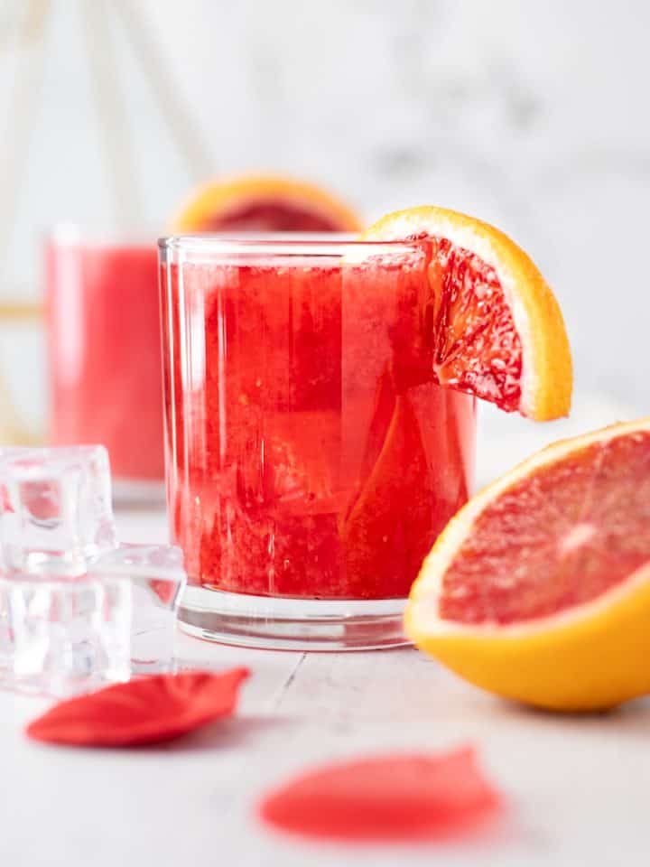 Glass of blood orange whisky sour, garnished with a slice of blood orange, with an orange slice ice cubes and rose petals in the foreground.
