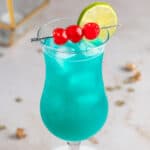 A blue hurricane drink garnished with maraschino cherries and a lime slice, on a marble table.