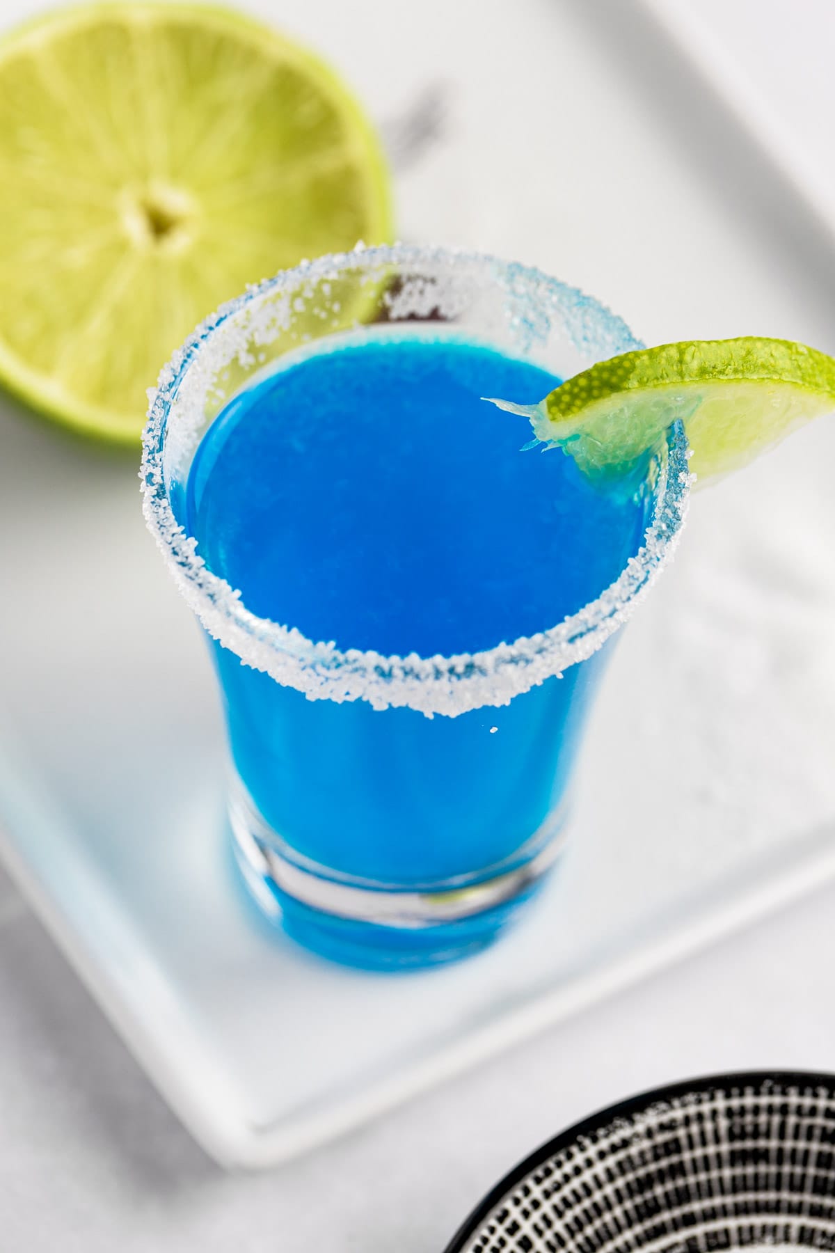 An overhead view of a blue kamikaze shot with a sugar rim, garnished with a slice of lime.