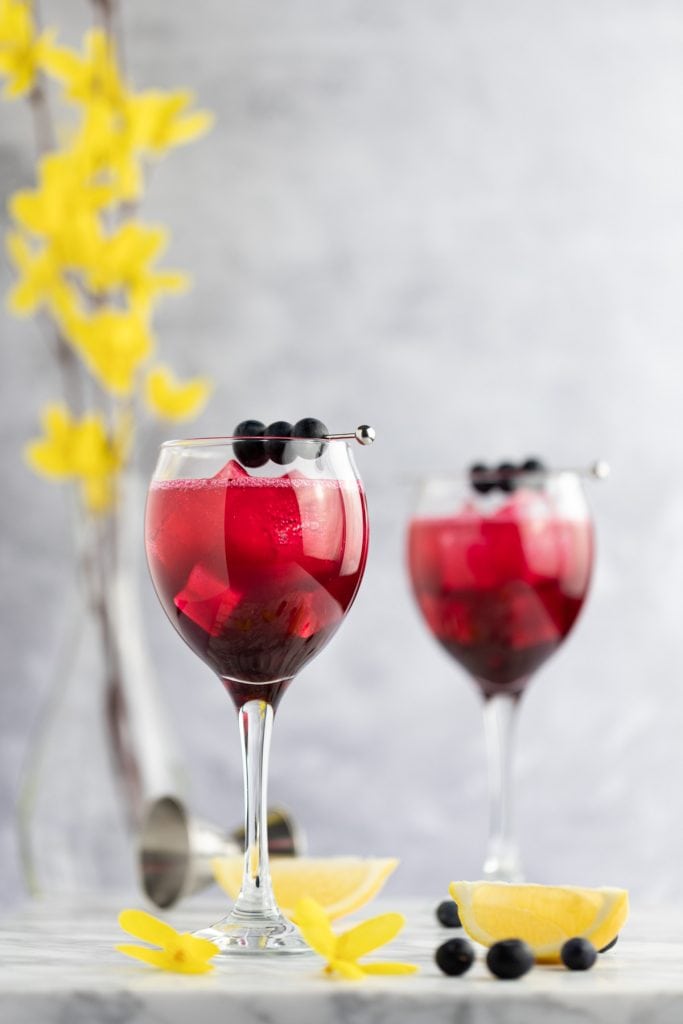 Two blueberry gin cocktails with blueberries as garnish, on a marble table next to lemon slices and yellow flowers.