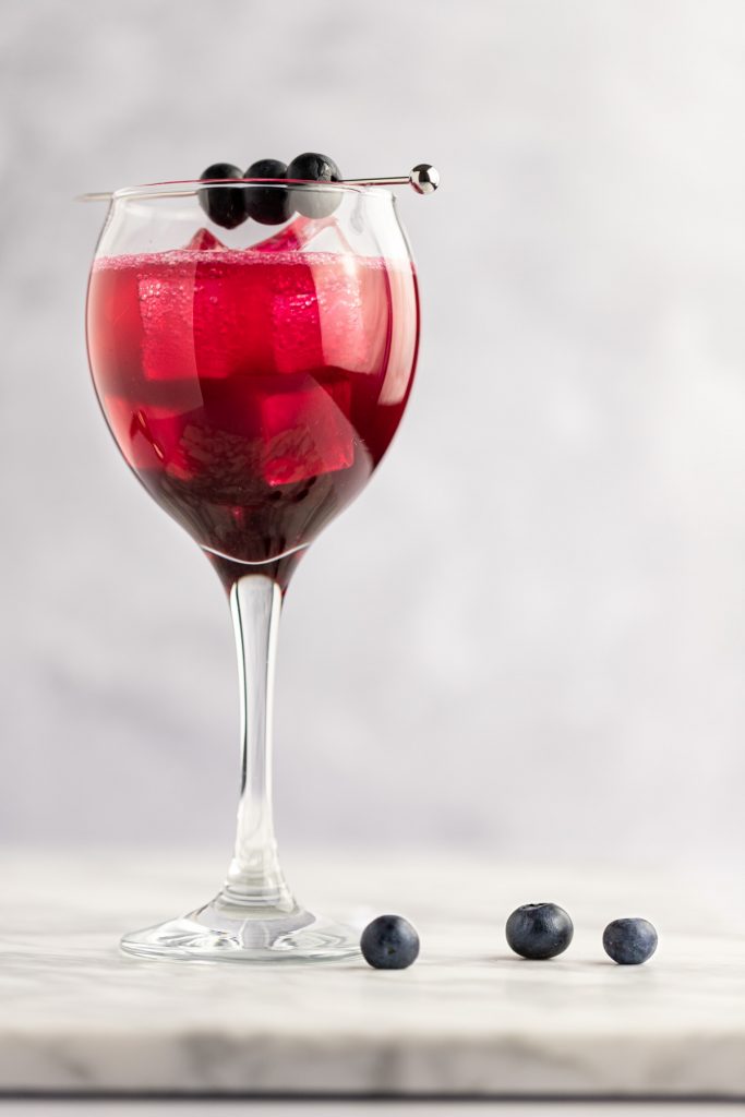 A single glass of blueberry cocktail on a white marble board, with three fresh blueberries beside it.