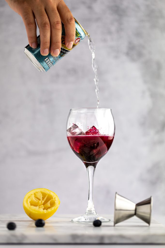 A hand holding a can of club soda being poured into a half filled blueberry cocktail with ice, next to a metal jigger, squeezed lemon and blueberries.