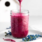 A spoon coated in blueberry glaze, dripping into a mason jar full of glaze, on a white background.