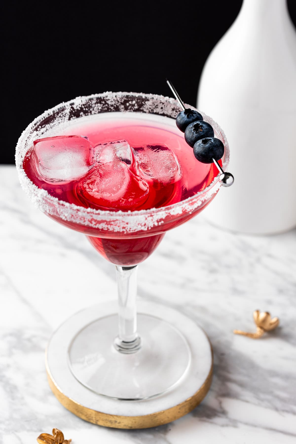 A blueberry lemonade margarita on a white and gold coaster with a black background.