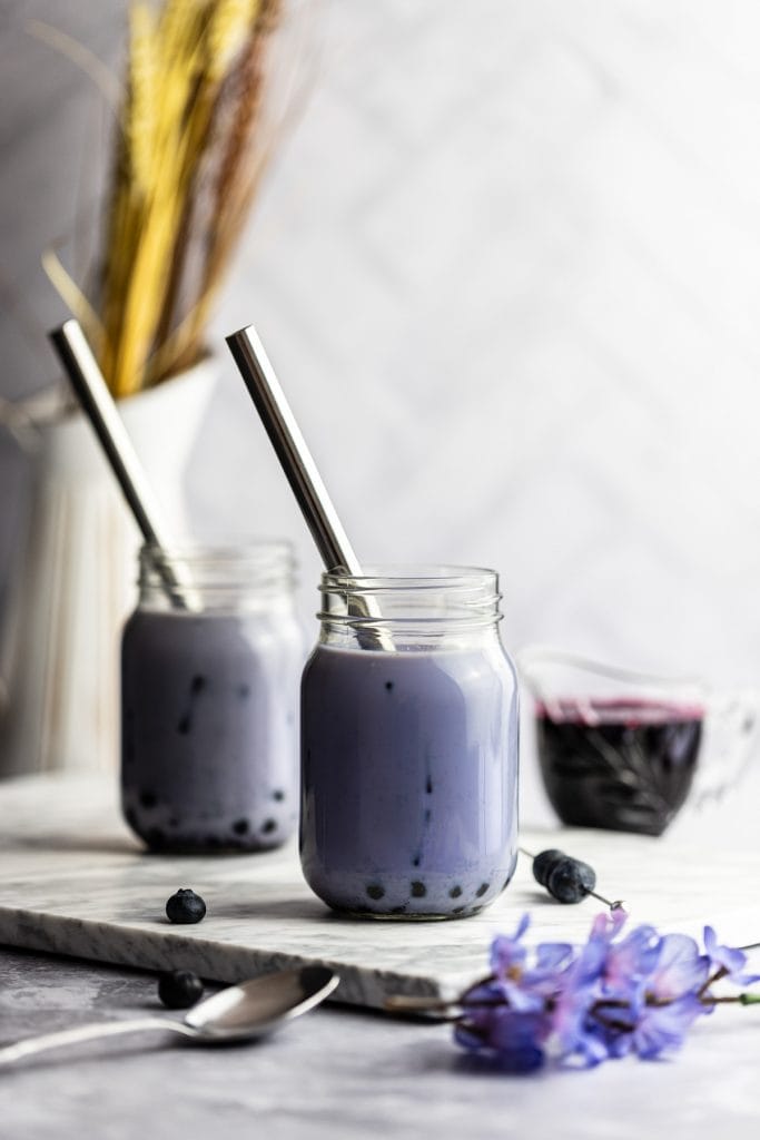 Two blueberry milk tea jars on a marble board, next to a jar of blueberry syrup, blueberries and a bunch of blue flowers.