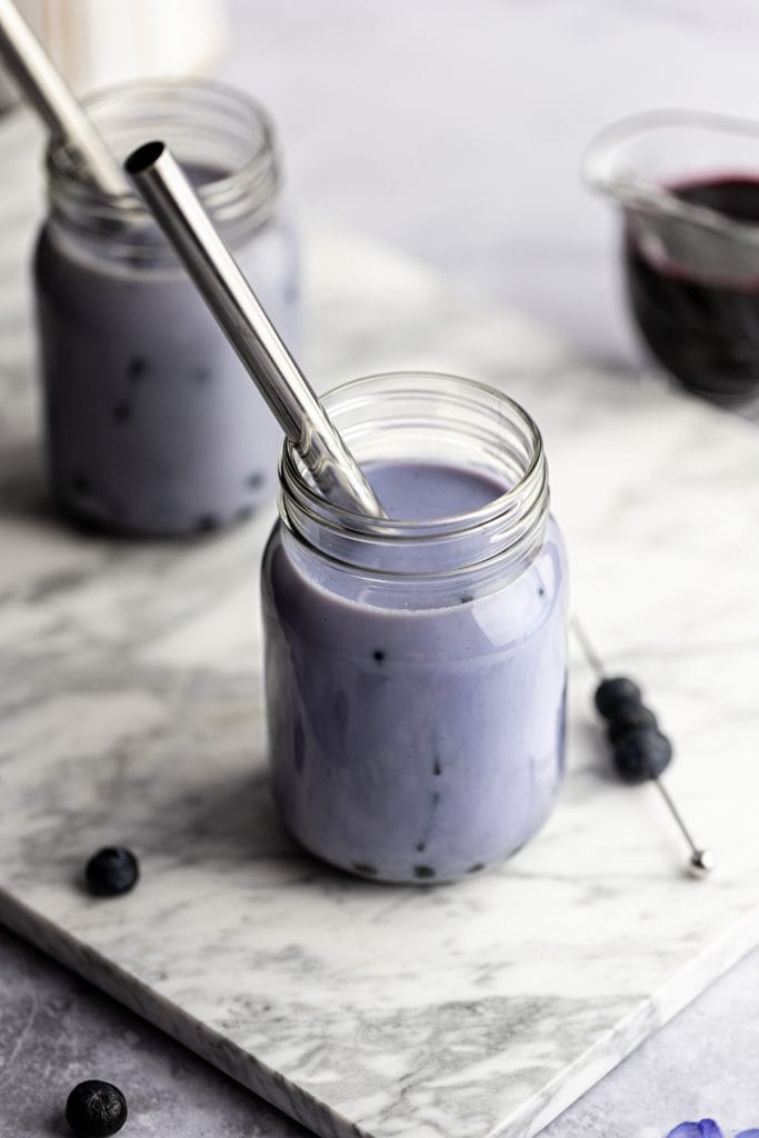 Overhead view of a glass of blue bubble tea on a grey marble board, next to a cocktail pick with three blueberries on it.