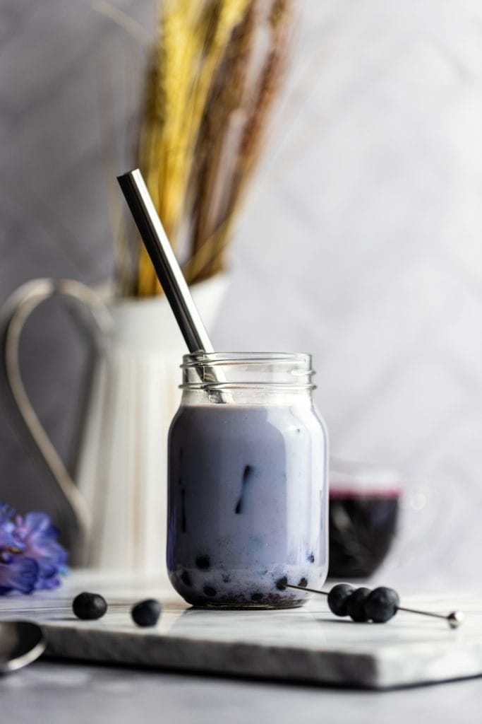 A shadowy photo of a blueberry bubble tea with boba, syrup and a white jug with wheat and purple flowers in the background.