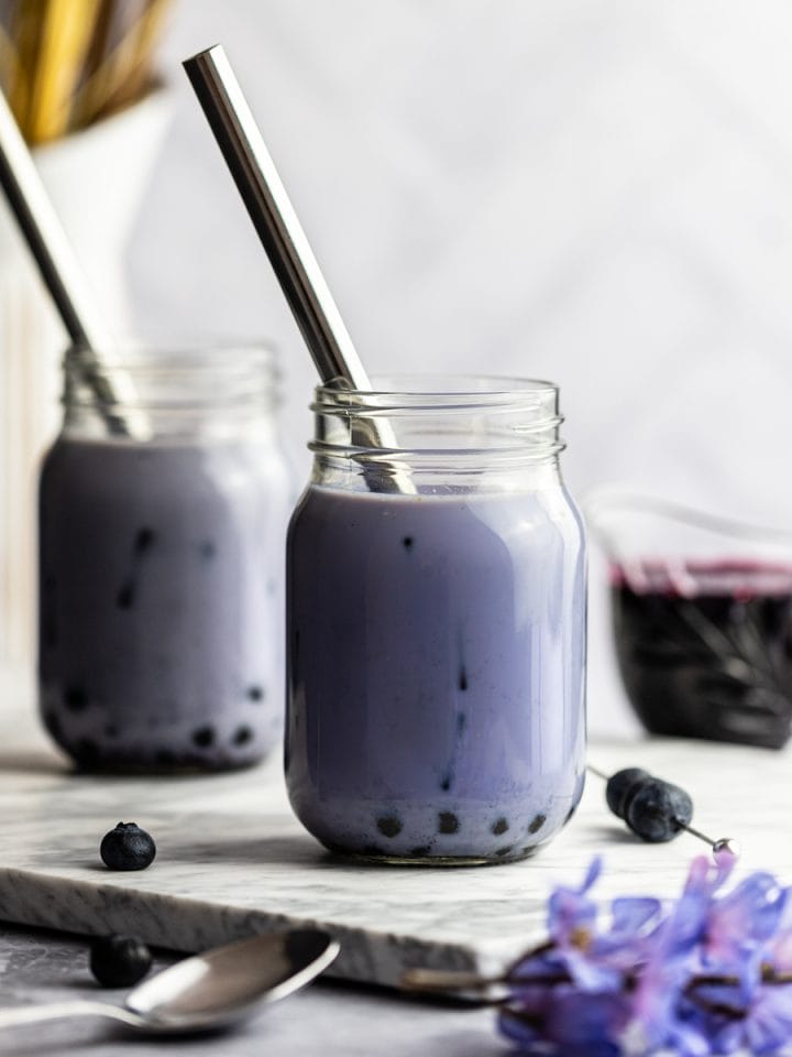 Two blueberry milk teas on a marble board next to blueberries, syrup and purple flowers.